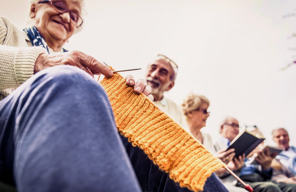 Photo of a group of seniors socializing. The focus of the photo is on a long piece of yellow knitting.
