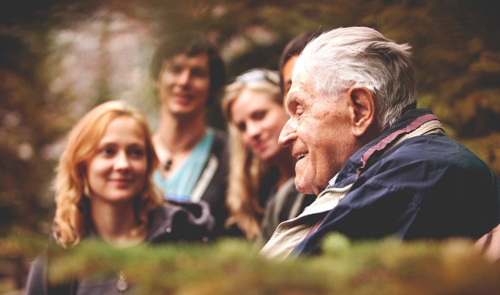 Photo of an elderly man in the foreground. He smiles and is surrounded by four young people.