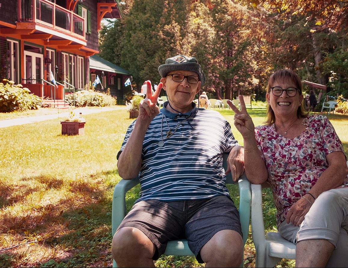 Photo of a senior couple in a sunny garden. They holding up two fingers in a "V" to make a peace signs.