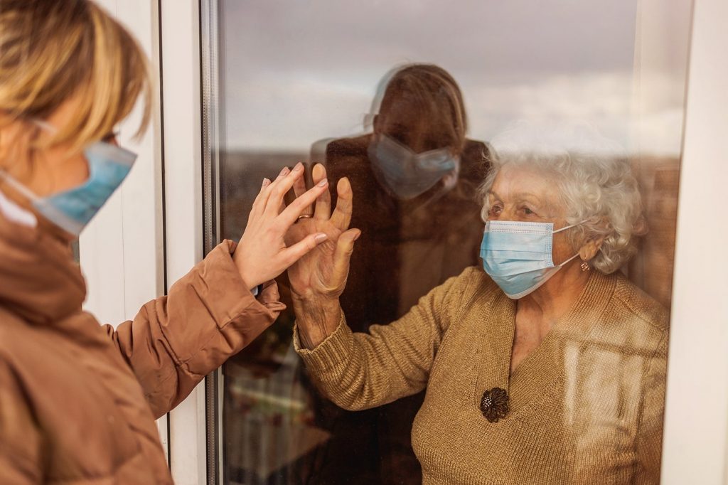 Photo of an elder and a young woman with disposable masks. They both put a hand against the window that separates them.