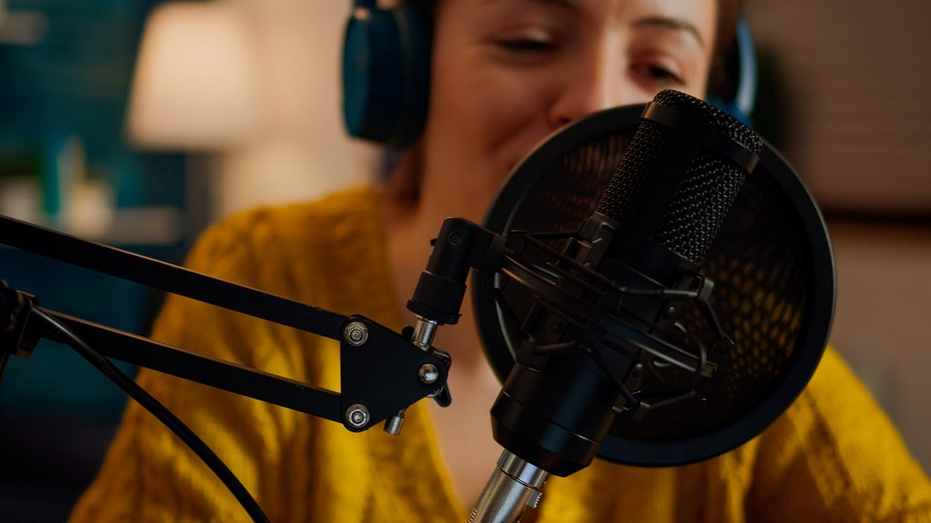 A photo of a young woman. The focus is on a studio microphone directly in front of her.