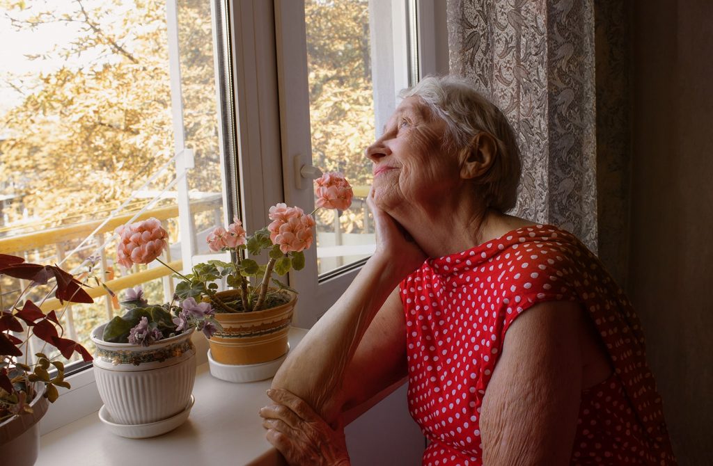 Photo d'une aînée qui regarde le ciel à travers sa fenêtre garnie de pots de fleurs.