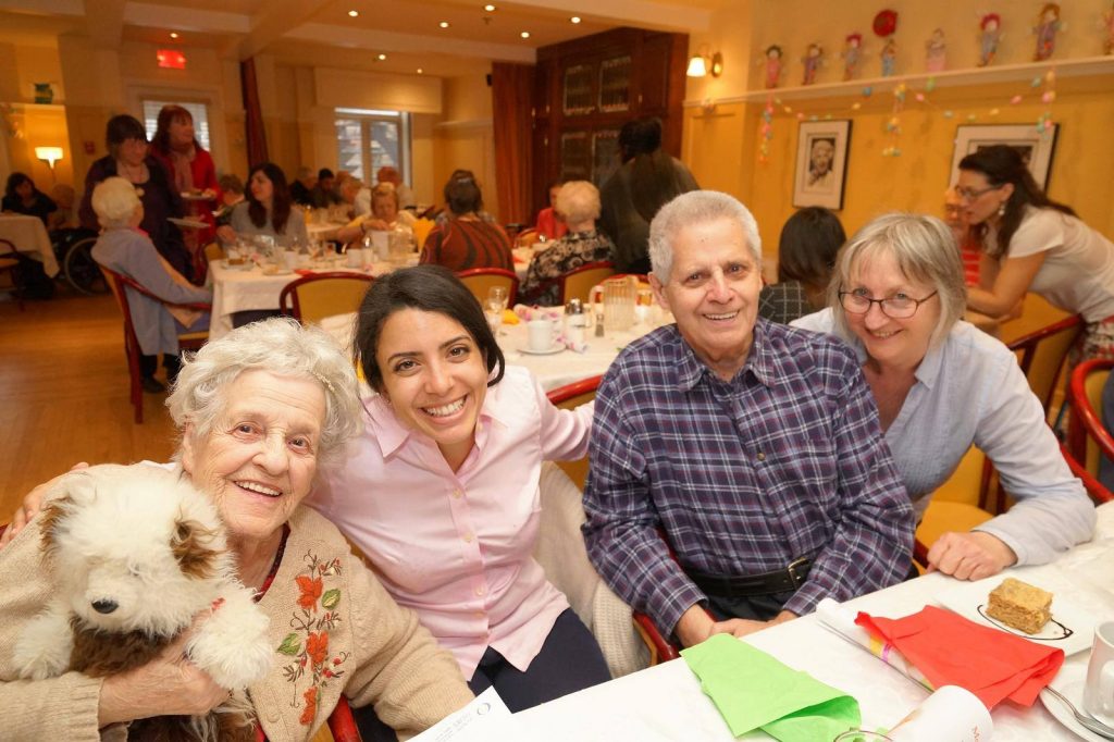Photo of a dinner party. The focus is on four people, including an elder with her dog in her arms.