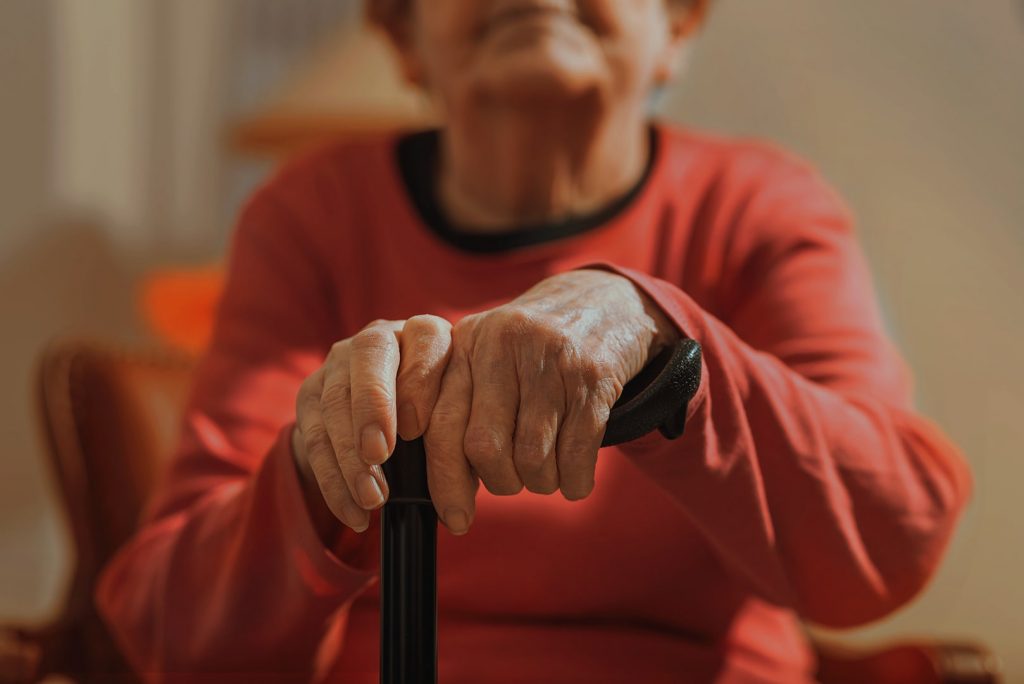 Photo of a seated elder. The focus is on her hands resting on her cane.