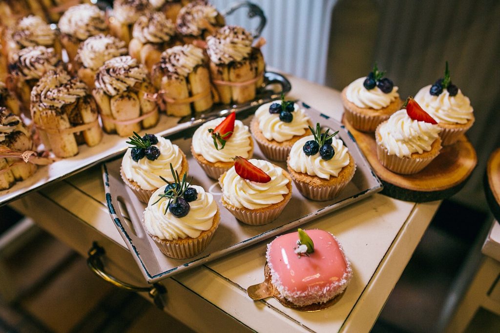 Photo of trays of individual servings of desserts. Cupcakes topped with glazed fruit, a small pink square rolled in coconut and mini charlottes.