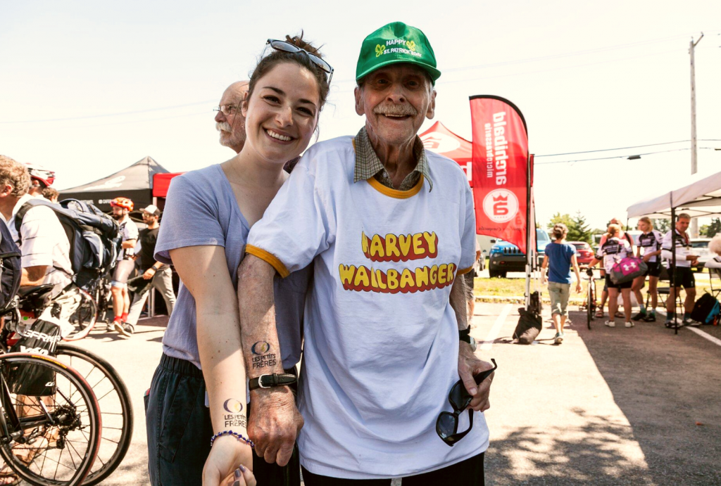 Photo of an elderly man at an event. He is accompanied by a young woman. They proudly show off their little brothers temporary tattoo.