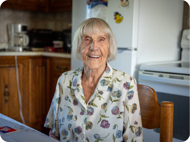 An older woman we support is seated in her kitchen, smiling and looking at the camera, illustrating a caring presence and a lived-in everyday setting.