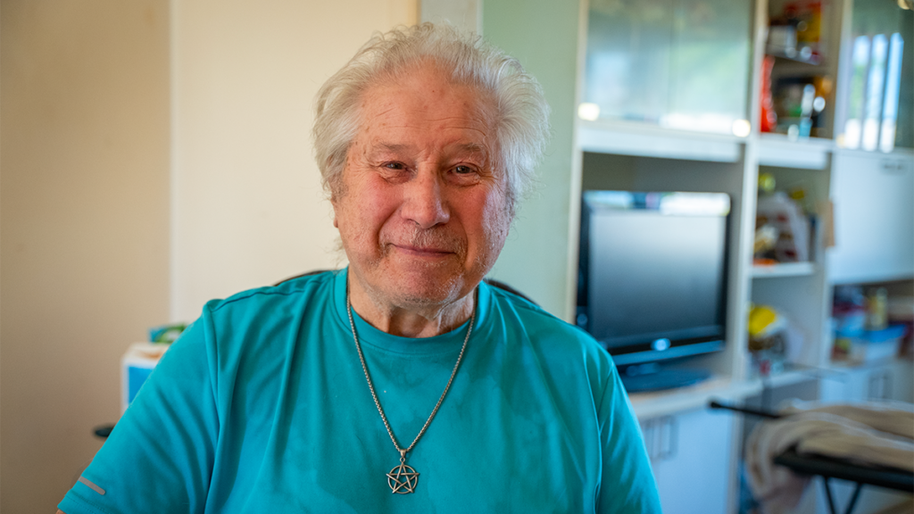 An older man we support is seated in his living room, smiling and looking at the camera.